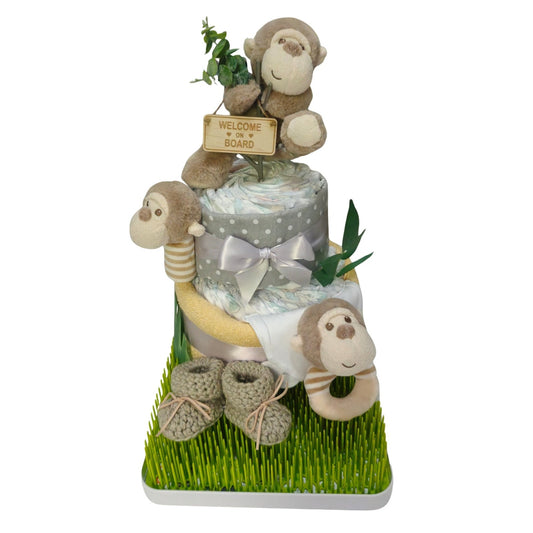 Cheeky monkey limited edition two-tier baby welcome nappy cake gift set featuring plush monkey rattles, knitted baby booties, and receiving blankets displayed on a green grass drying rack base.