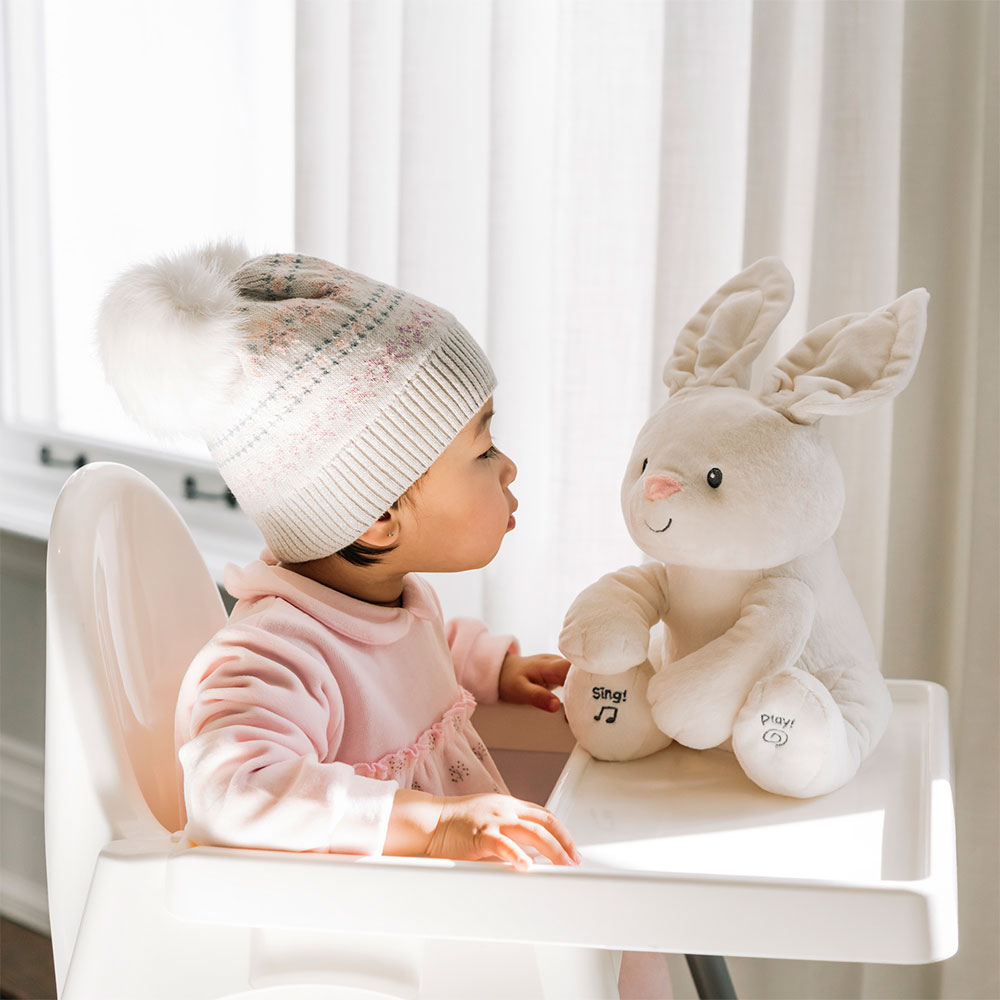 Toddler sitting beside GUND Flora Bunny animated plush toy on a high chair tray