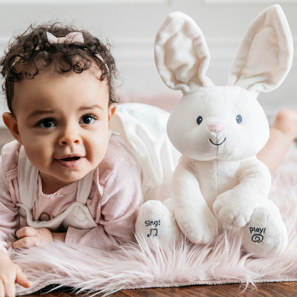 Baby lying beside GUND Flora Bunny animated plush toy on a soft pink blanket