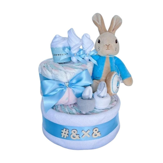 Two-tier nappy cake with a Peter Rabbit plush, blue ribbons, baby socks, and wooden "#&X&" letters.