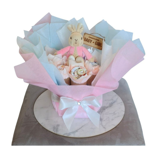 Front view of a baby girl bouquet with a pink-jacketed plush rabbit, rolled pink bibs, and a "Baby Girl" sign.