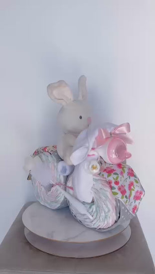 360-degree video of a baby girl tricycle diaper cake gift set on a rotating display. Features a white plush bunny toy rider, floral blankets, a pink baby bottle, and a wooden 'Congratulations Baby Girl' sign