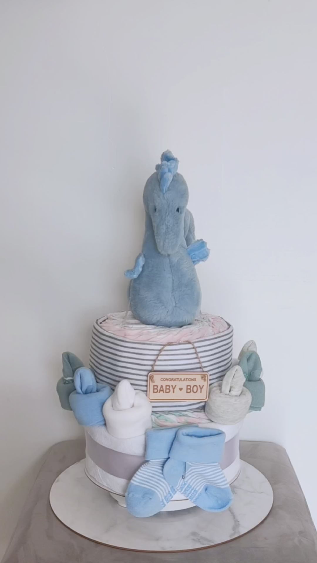 360-degree rotating view of a blue seahorse 1-tier baby boy nappy cake gift set, featuring a plush seahorse toy, receiving blanket, rolled infant nappies, baby booties, and a wooden Congratulations Baby Boy sign.