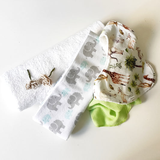 Flat lay of Woodie unisex baby owl nappy cake components featuring a safari animal print baby bib, grey receiving blanket, towel, and decorative fabric flowers.